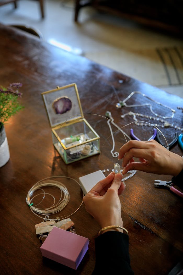 Person crafting jewelry on a table using wires and tools, featuring a glass container and accessories.